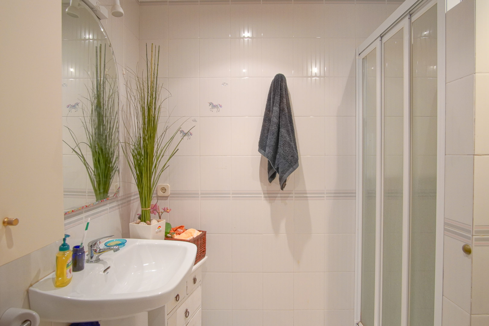 Baño funcional con paredes alicatadas en blanco, lavabo de porcelana, espejo de arco y zona de ducha con mampara corredera blanca.