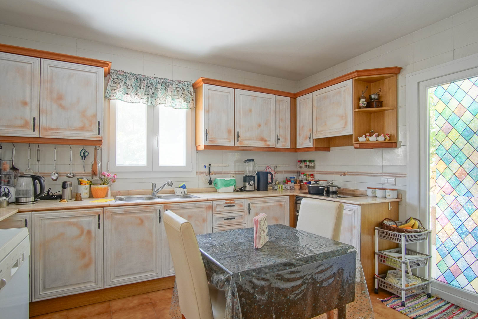 Cocina independiente de estilo rústico con armarios de acabado decapado, encimera en L y zona de comedor. Incluye una luminosa ventana y acceso exterior mediante una puerta con vidriera decorativa.