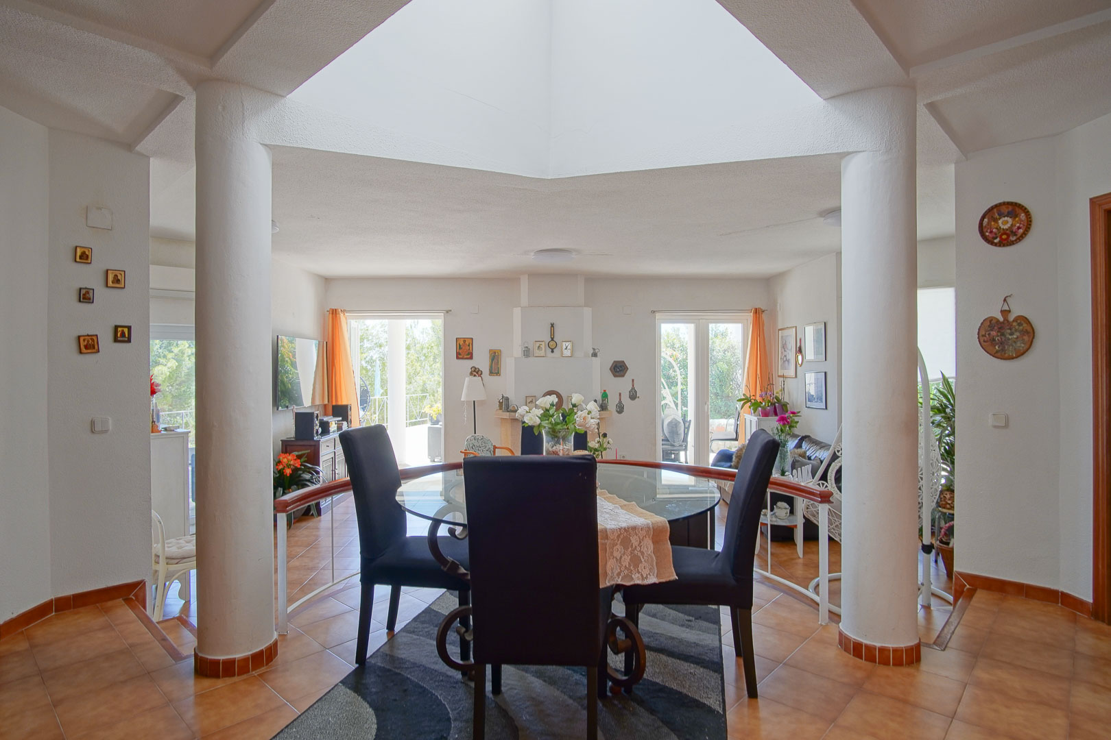 Comedor luminoso bajo un lucernario central, enmarcado por columnas blancas y suelos de terracota que conectan con el salón y la chimenea de fondo.