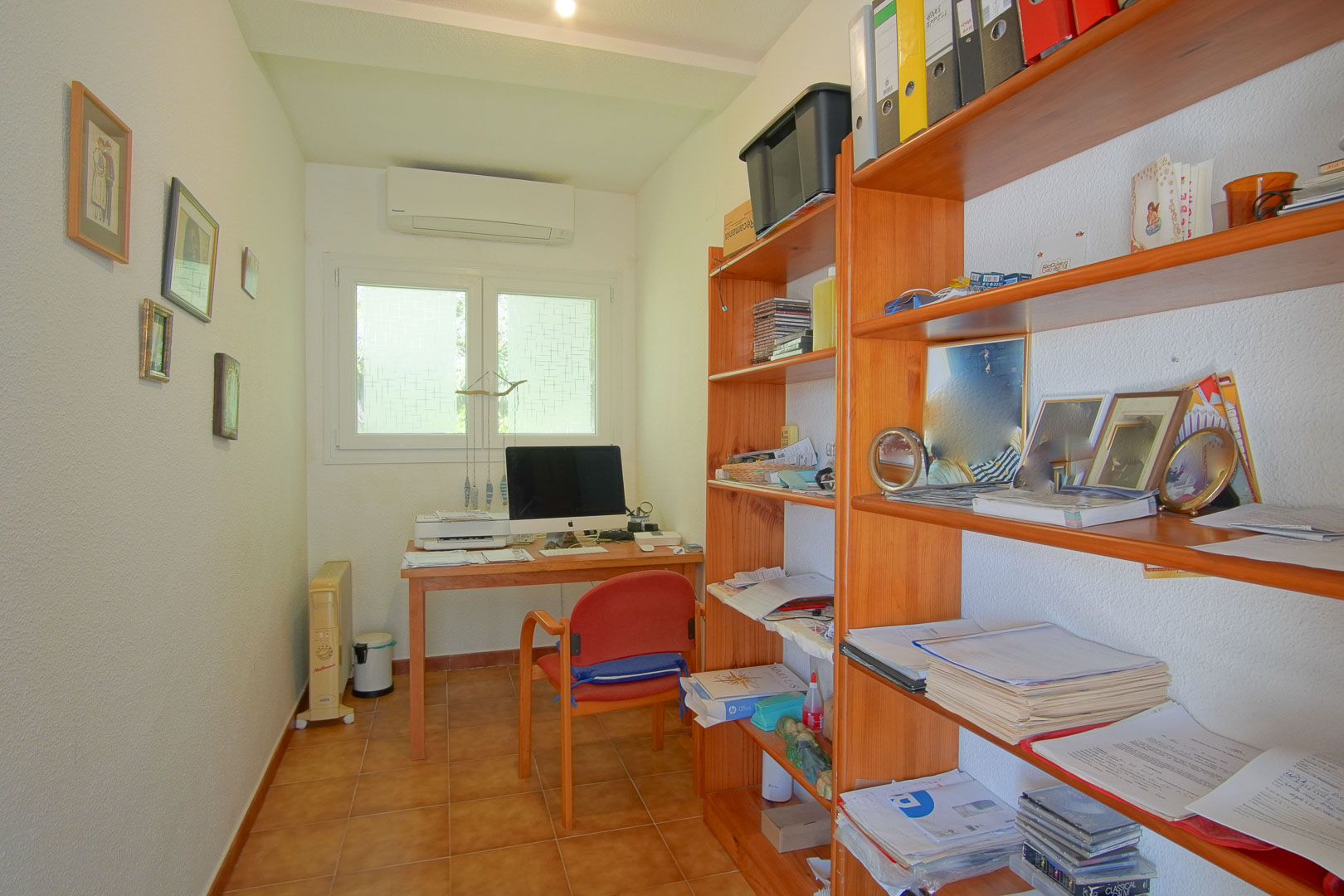 Despacho funcional con escritorio de madera, estanterías integradas, suelos de gres y aire acondicionado, ideal para el teletrabajo con luz natural.