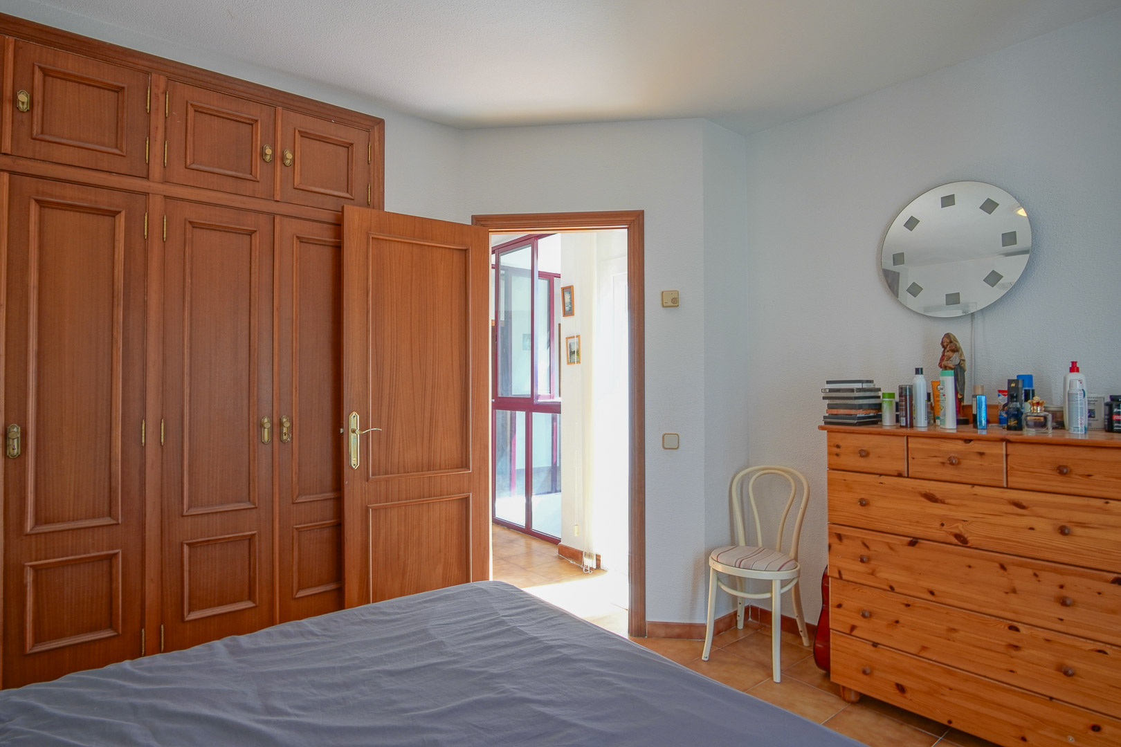 Dormitorio con amplios armarios empotrados de madera, cómoda a juego y suelos de gres. La estancia conecta con un pasillo luminoso que resalta la calidez de la carpintería clásica.