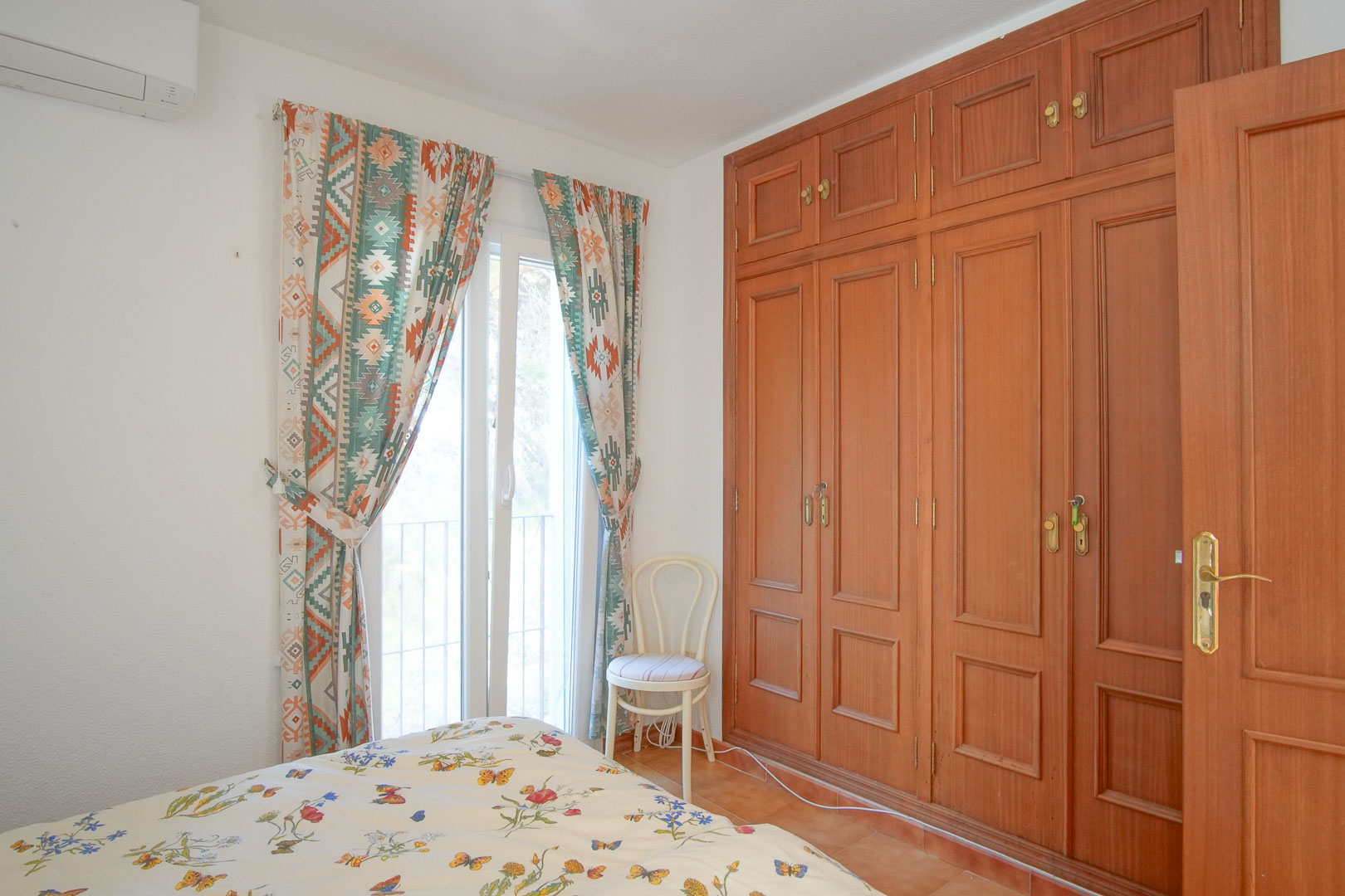 Dormitorio luminoso con armarios empotrados de madera de suelo a techo, aire acondicionado y acceso directo al exterior a través de un ventanal con cortinas decorativas.