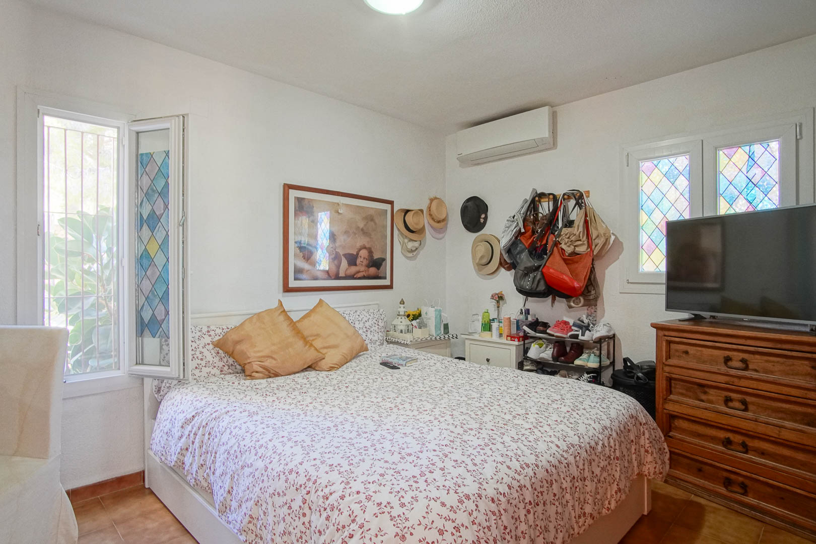 Dormitorio luminoso con suelos de gres, aire acondicionado split y ventanas decoradas con vidrieras. Incluye una cómoda de madera clásica y abundante luz natural.