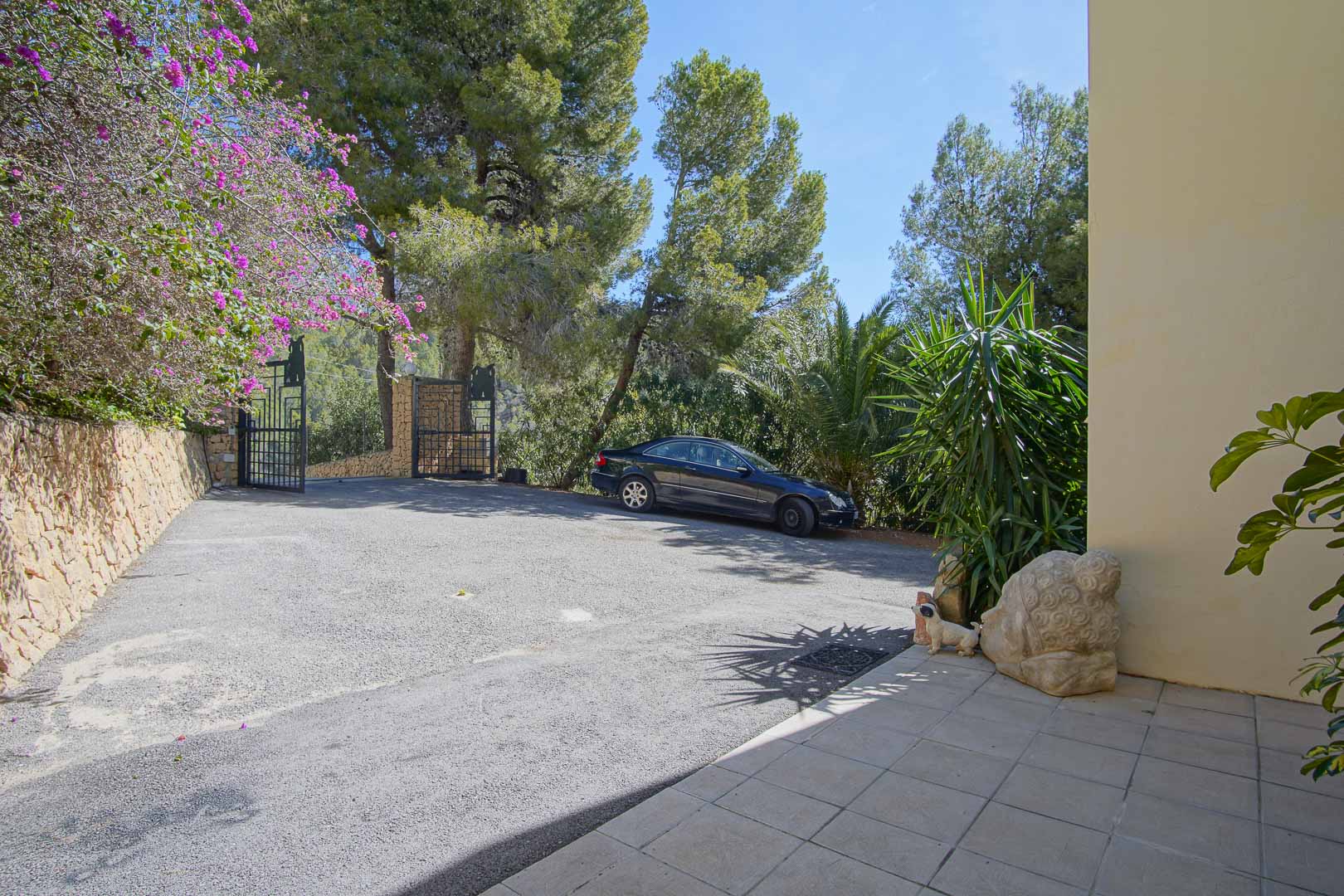 Entrada principal con portón de hierro, amplia zona de aparcamiento pavimentada y muros de piedra natural rodeados de pinos y vegetación autóctona.