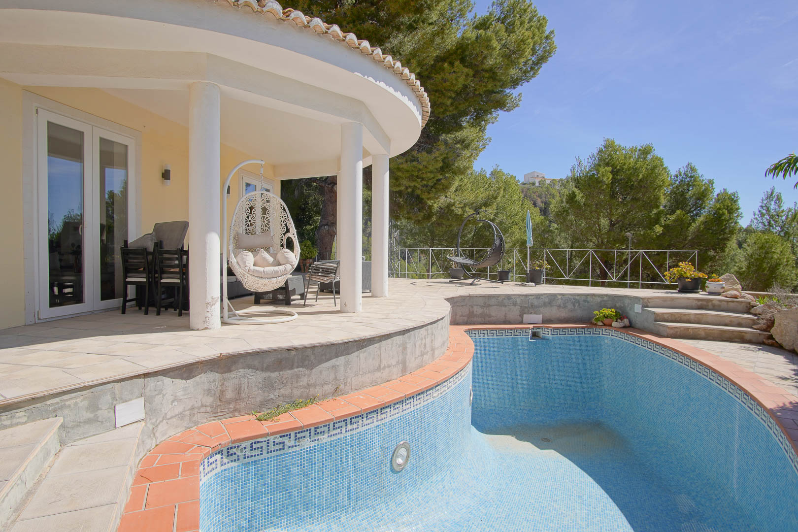 Porche cubierto de diseño curvo con columnas blancas y zona de estar, situado junto a una piscina de gresite azul rodeada de pinos mediterráneos.