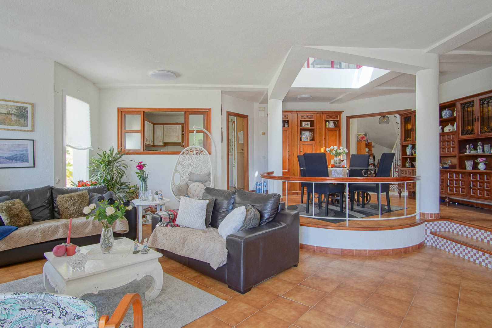 Salón de planta abierta con suelos cerámicos, sofás de cuero y un comedor elevado sobre plataforma circular con barandilla de madera y claraboya superior.