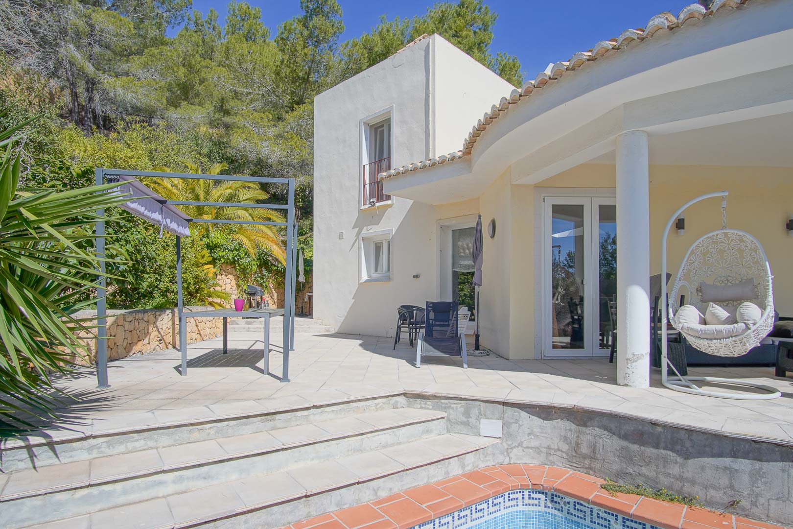 Terraza exterior con piscina, zona de comedor bajo pérgola y porche cubierto con sillón colgante, rodeada de vegetación mediterránea y pinos.