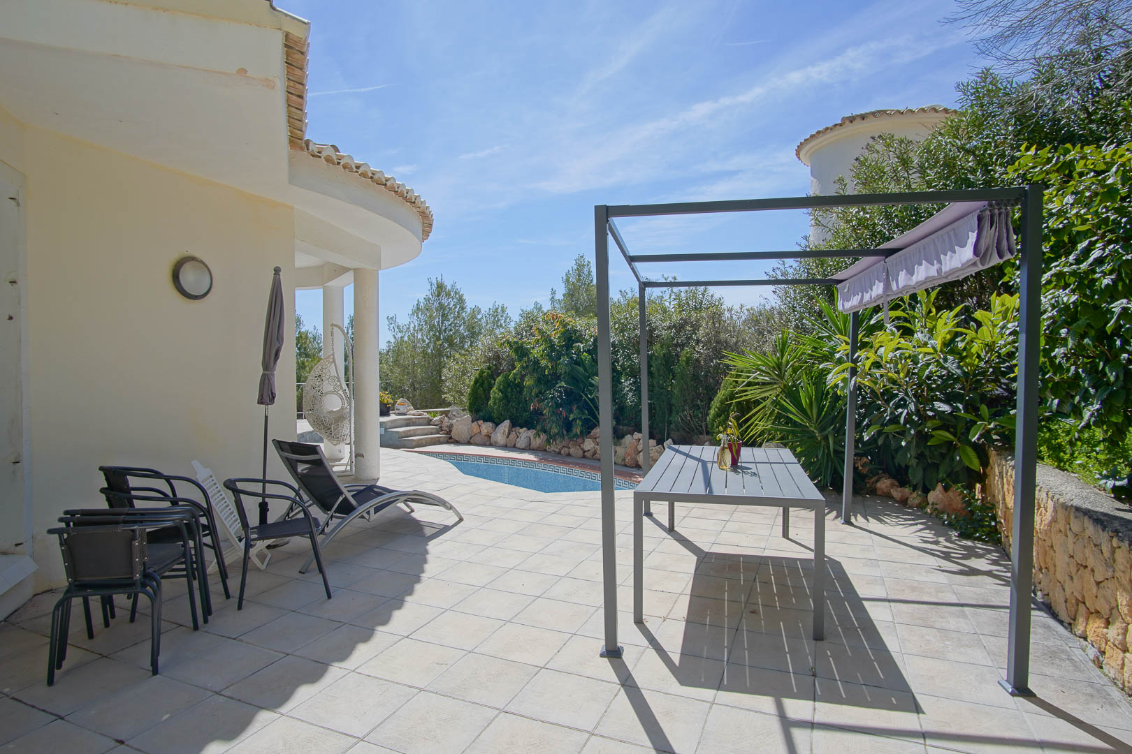 Terraza exterior con suelo cerámico, zona de comedor bajo pérgola metálica y vistas a la piscina privada rodeada de vegetación mediterránea.