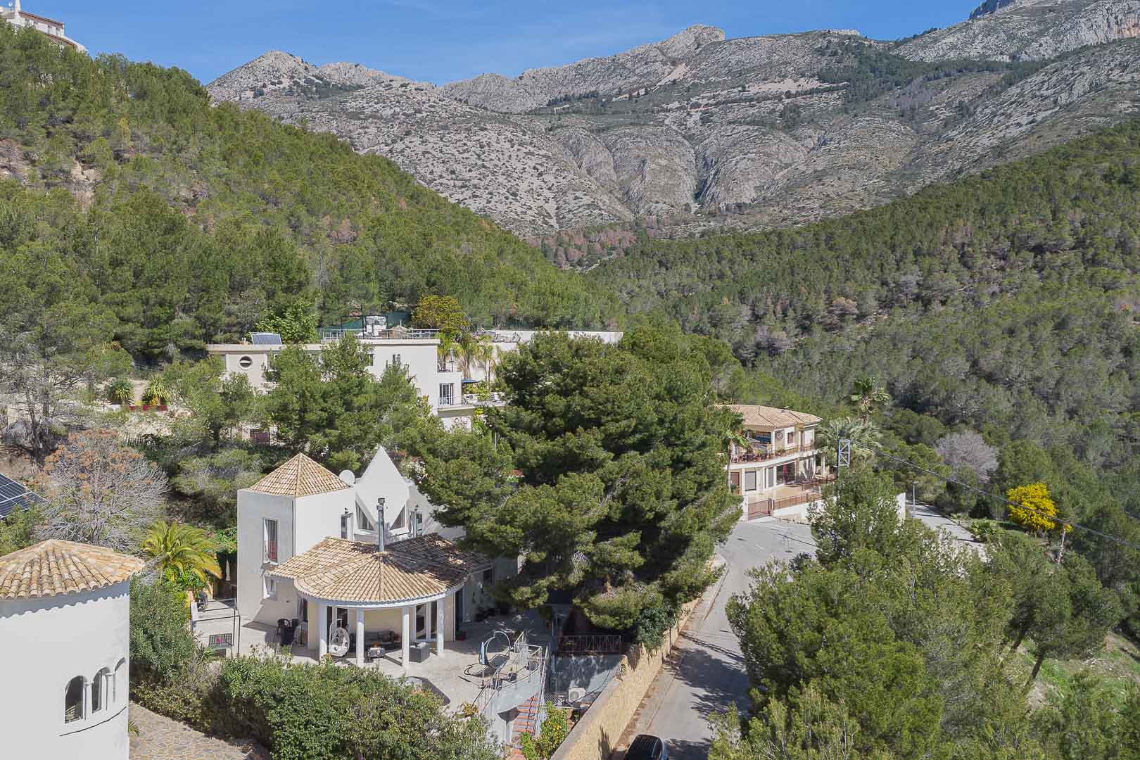 Vista aérea de la villa destacando su arquitectura mediterránea con techos de teja, rodeada por un denso bosque de pinos y las majestuosas montañas de Altea.