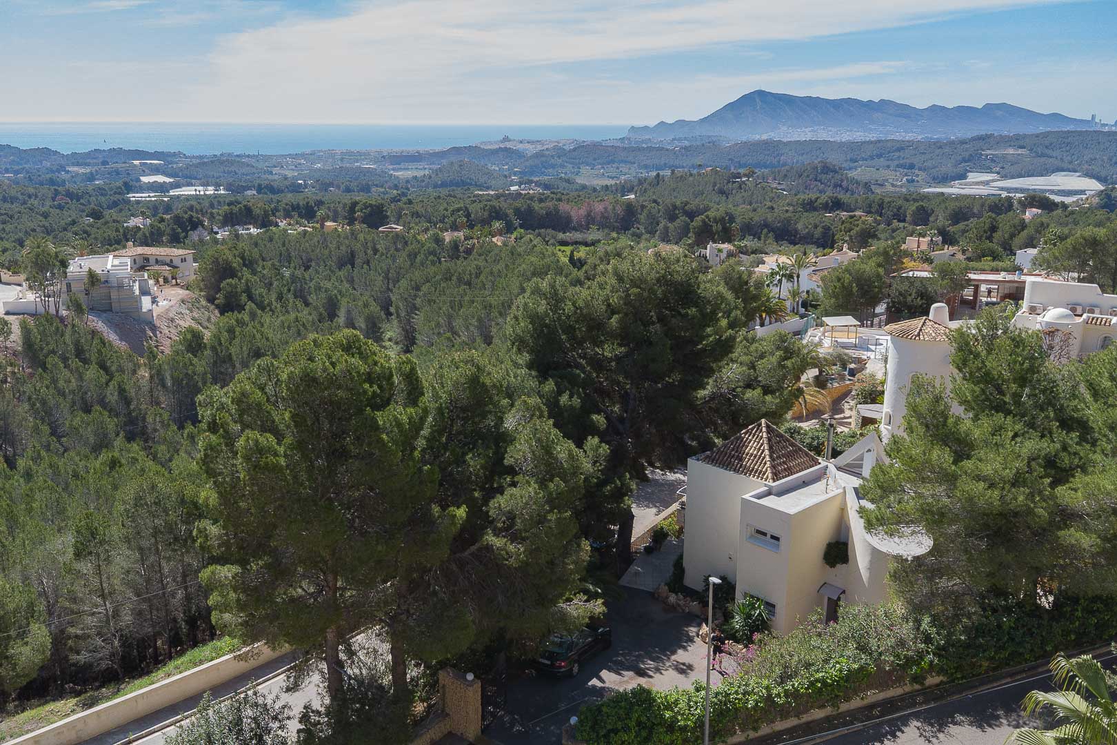 Vista aérea panorámica que muestra la villa integrada en un entorno de pinos, con vistas despejadas al mar Mediterráneo y a las montañas bajo un cielo despejado.