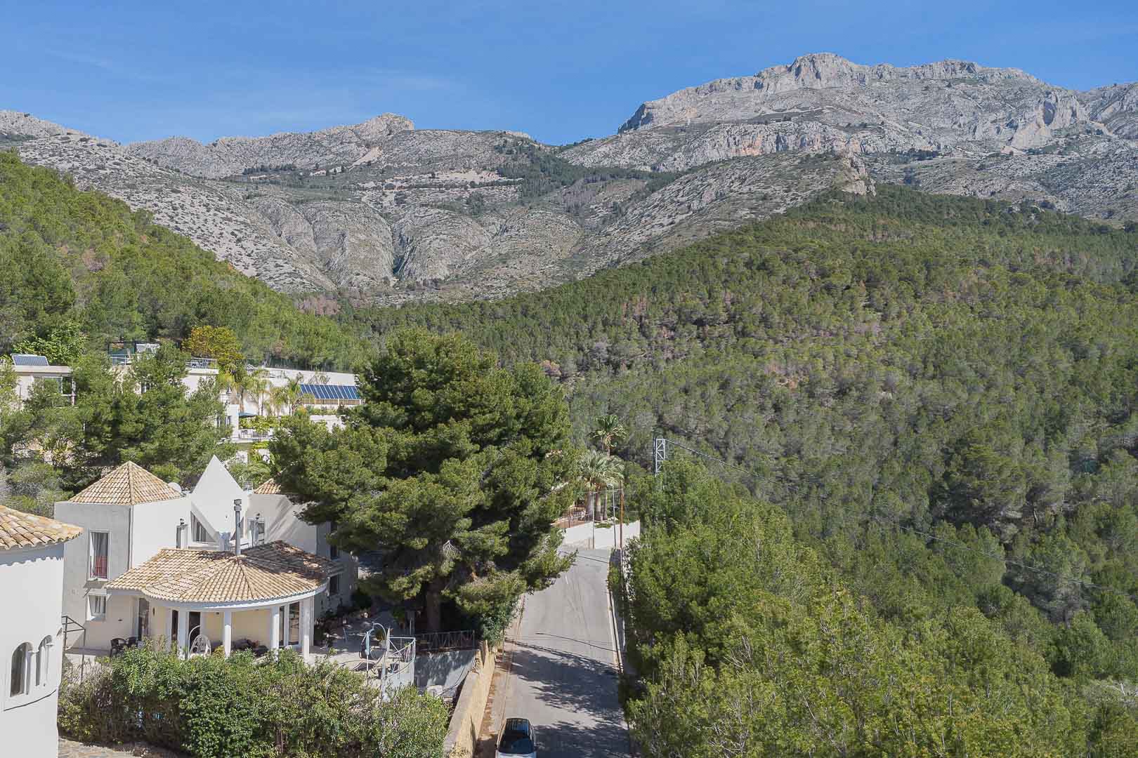 Vista panorámica de la villa integrada en la naturaleza, destacando su arquitectura mediterránea frente a la frondosa pinada y las imponentes montañas de Altea.