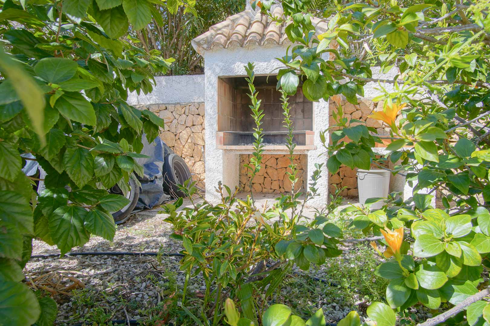 Zona de barbacoa de obra con tejado de teja cerámica, integrada en un entorno de jardín mediterráneo con muros de piedra natural.