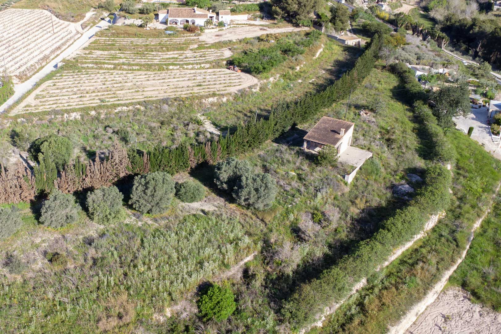 Vista aérea de la parcela de 10.920 m2 en Teulada, mostrando el terreno abancalado con vegetación mediterránea y una construcción rural existente.