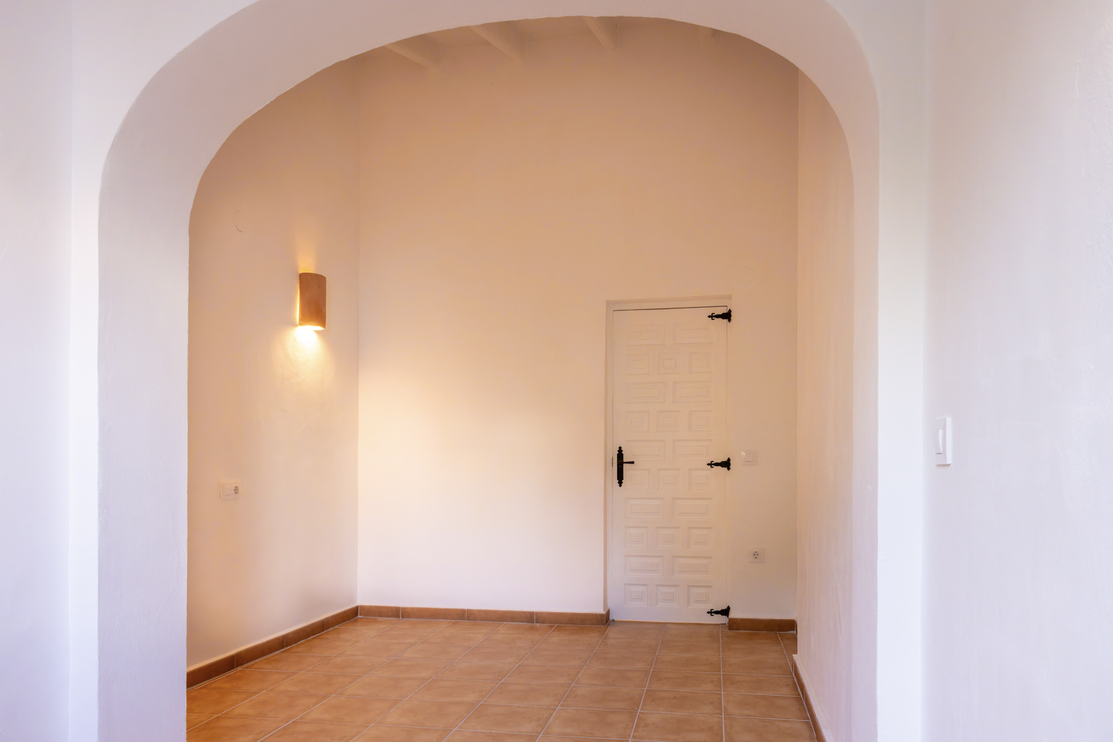 Entrada luminosa con arco, baldosa rústica y puerta blanca con herrajes negros. Aplique cilíndrico de pared.