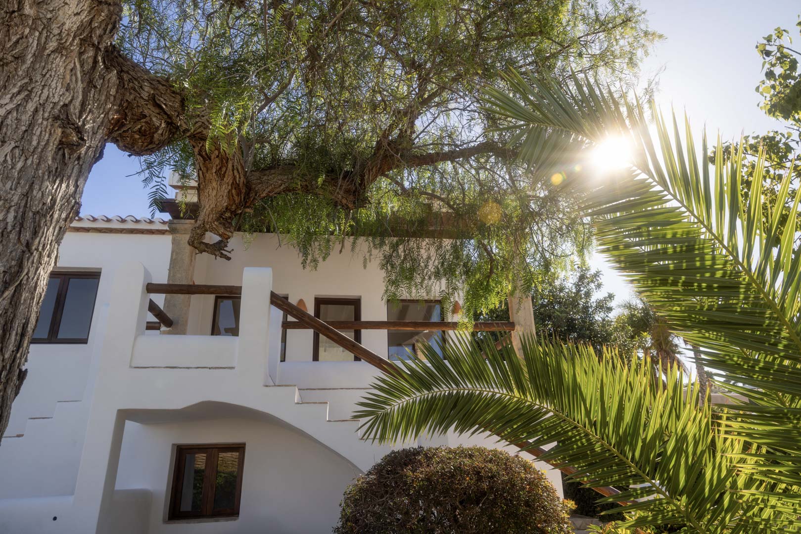Fachada blanca con balcón de madera y vegetación exuberante, destacando la arquitectura mediterránea.