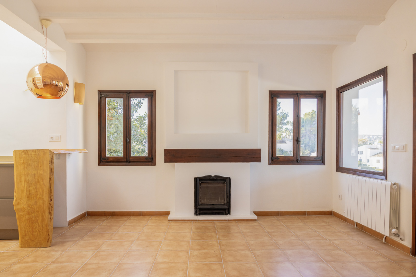 Salón luminoso con chimenea rústica, ventanas de madera y suelo de baldosas. Diseño mediterráneo con toques modernos.