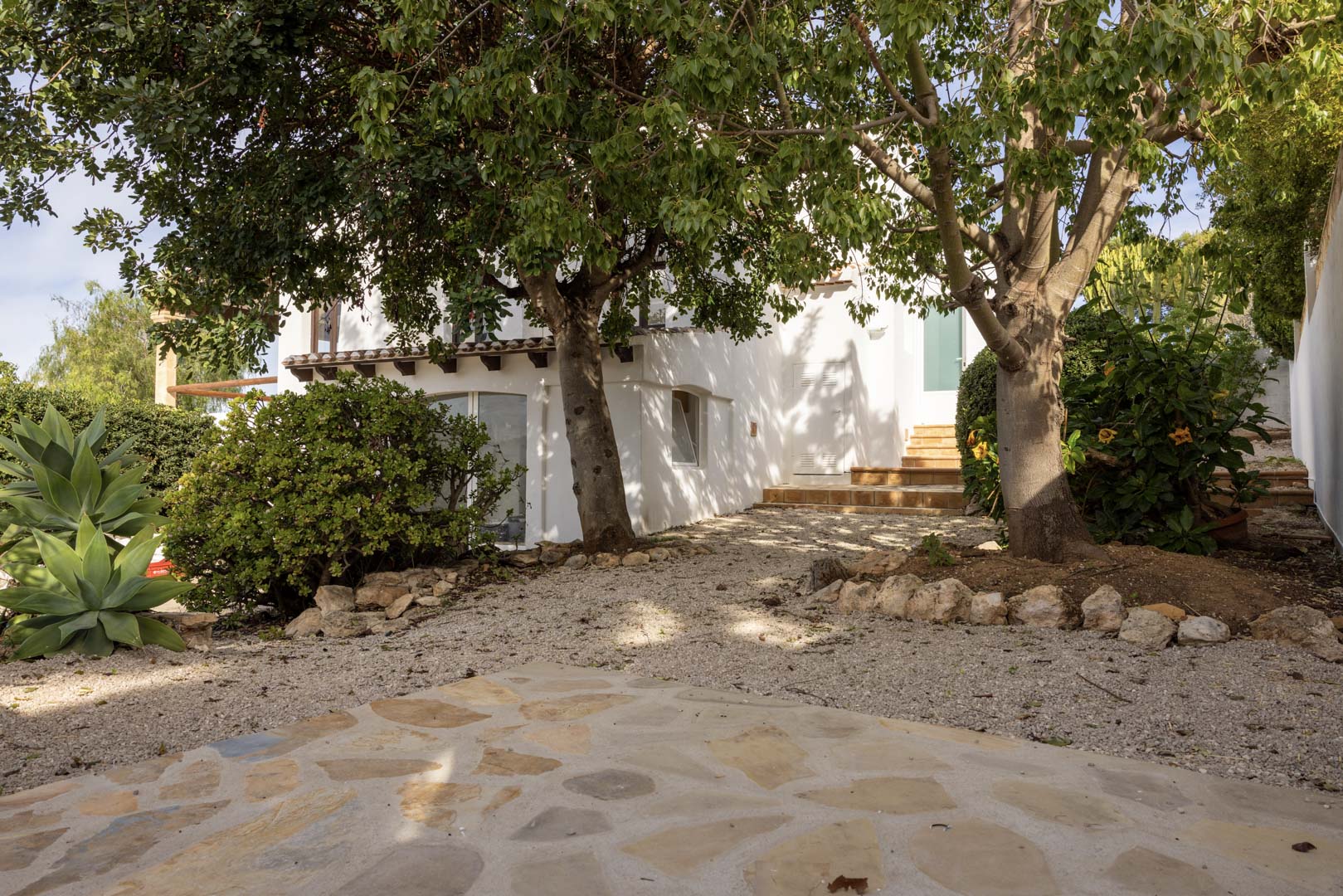 Vista exterior de la villa con jardín de grava y acceso a la entrada. Vegetación madura y fachada blanca.