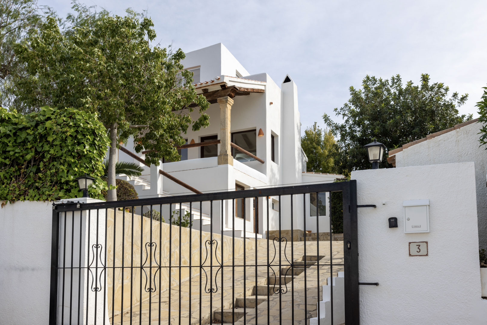 Vista exterior de la villa con puerta de hierro forjado y fachada blanca. Detalles de piedra y vegetación exuberante.