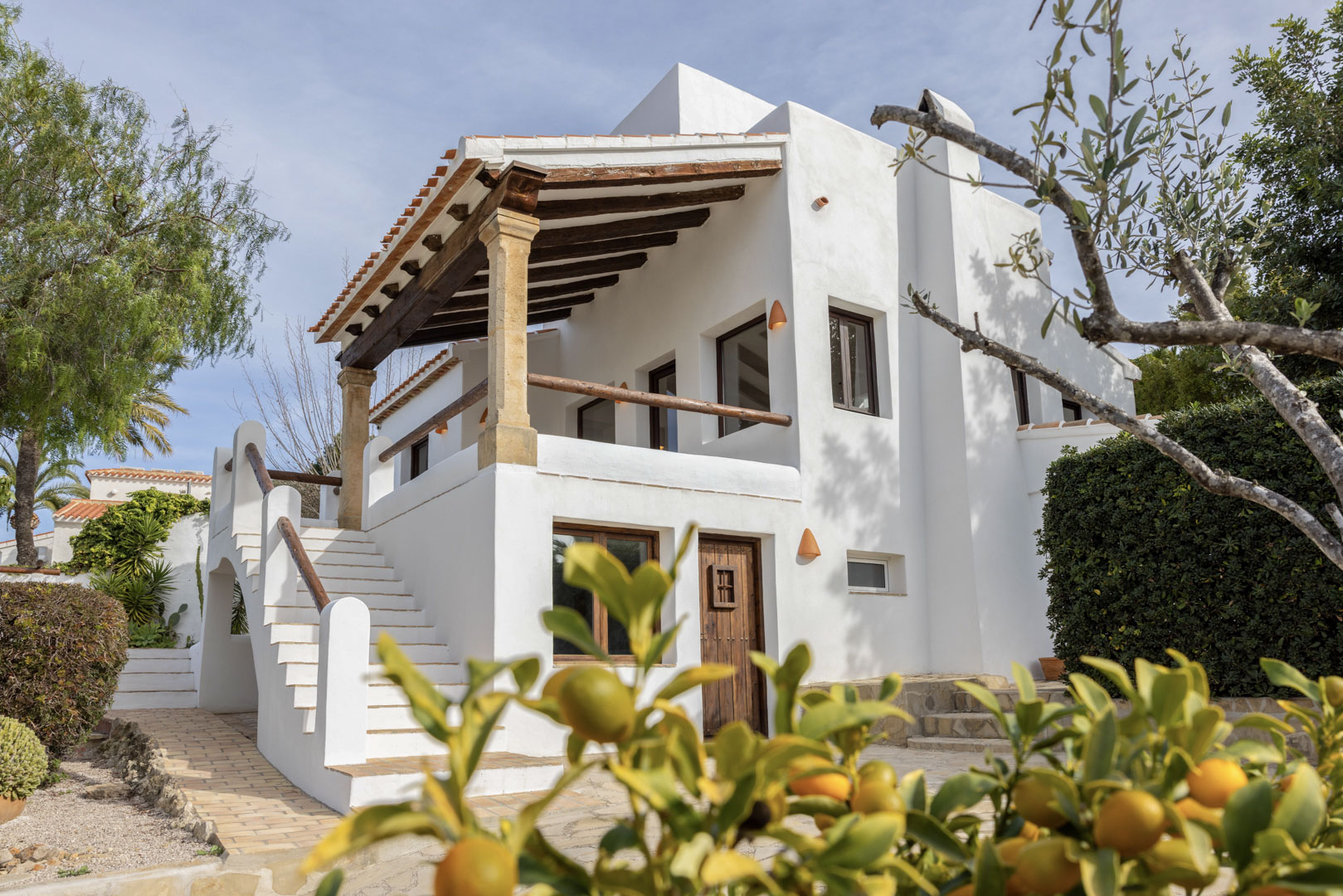 Vista exterior de la villa en Jávea, destacando la fachada blanca con terraza cubierta de vigas de madera y acceso por escalera.