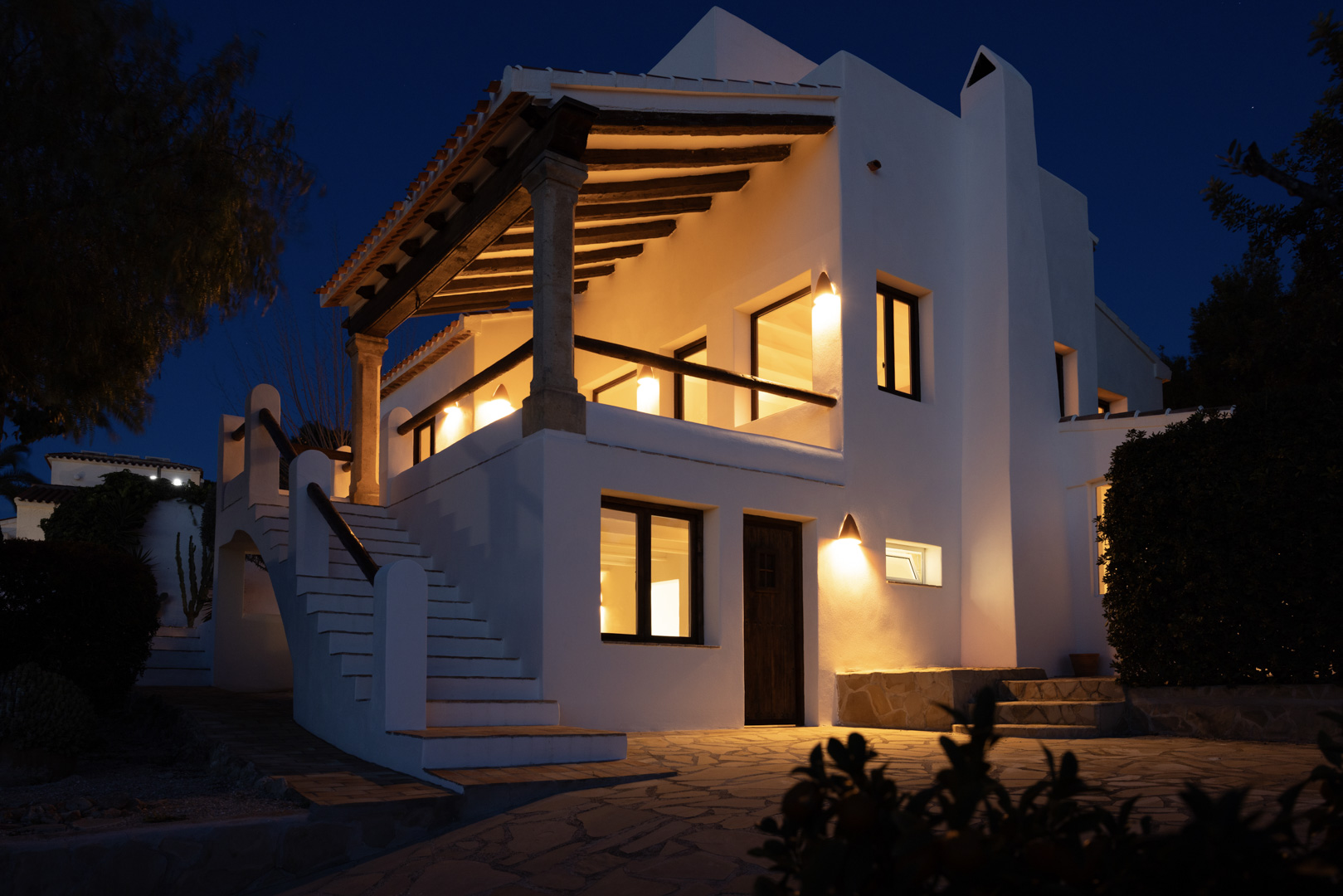 Vista nocturna de la villa con terraza cubierta, iluminada y escalera exterior de piedra.