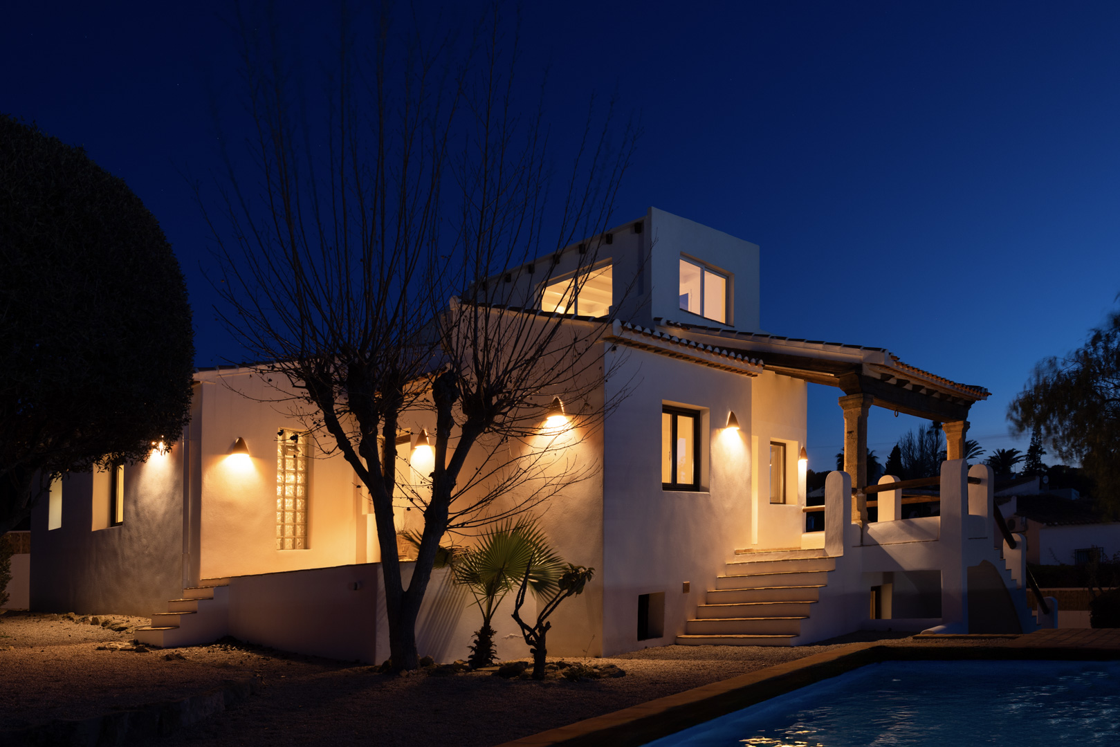 Vista nocturna de la villa iluminada con piscina reflectante y fachada blanca tradicional.