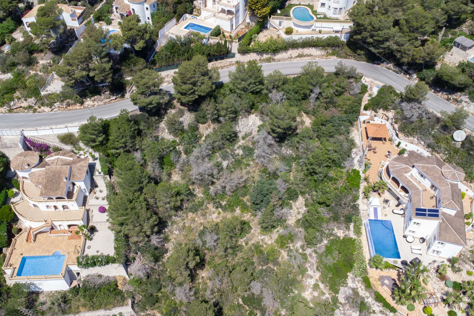 Vista aérea cenital de la parcela inclinada con vegetación mediterránea autóctona, situada entre villas residenciales con piscina en la zona de Costa Nova.