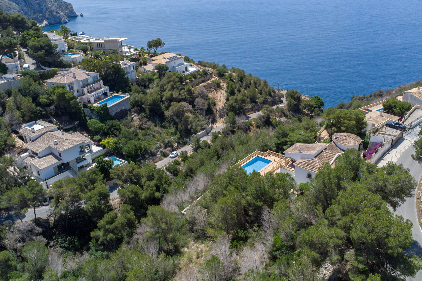 Vista aérea de la costa de Jávea que muestra villas mediterráneas rodeadas de pinos y el mar azul al fondo, destacando la proximidad de las parcelas al litoral.