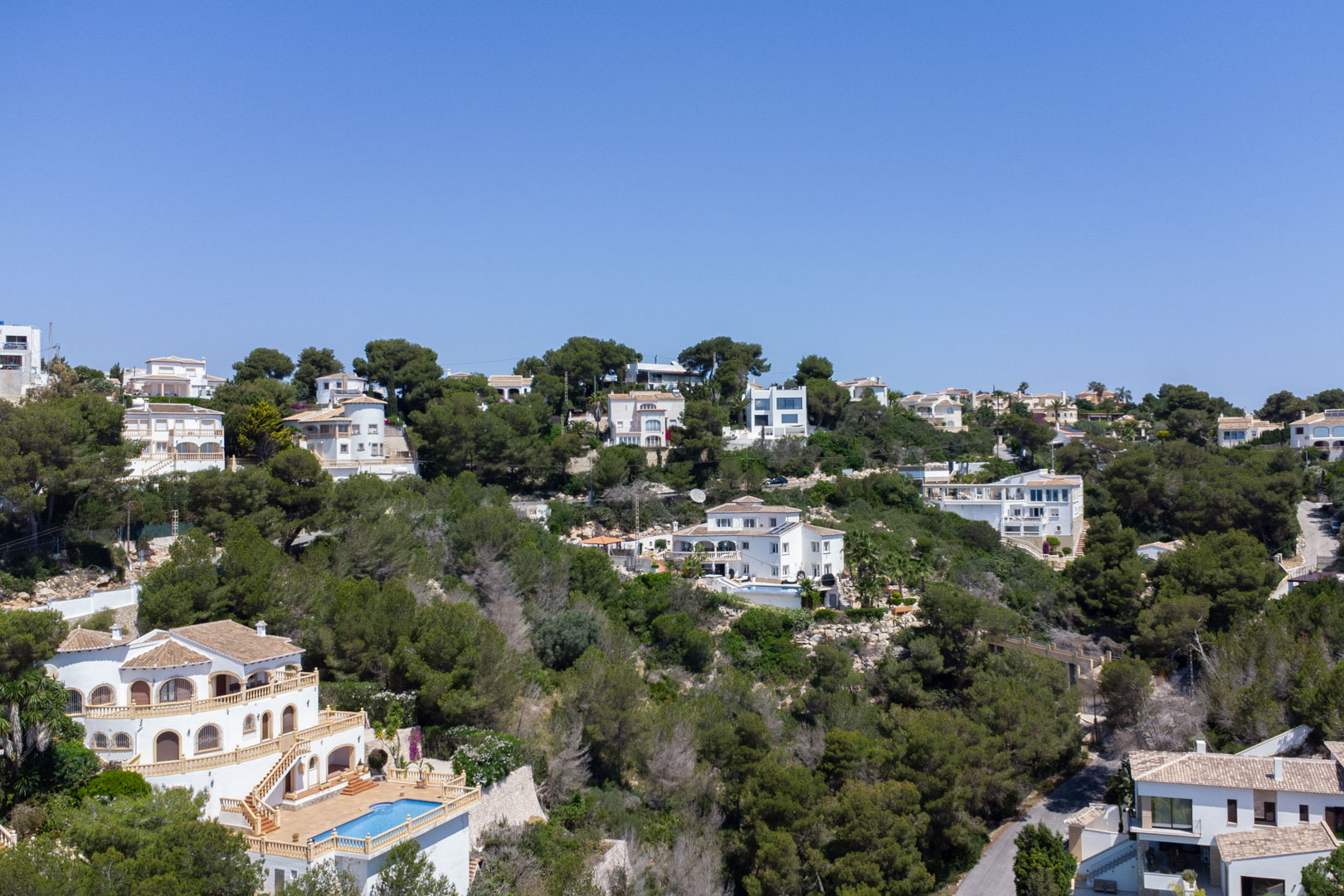 Vista aérea de la urbanización Costa Nova en Jávea, mostrando villas mediterráneas con piscinas integradas en una ladera arbolada bajo un cielo despejado.