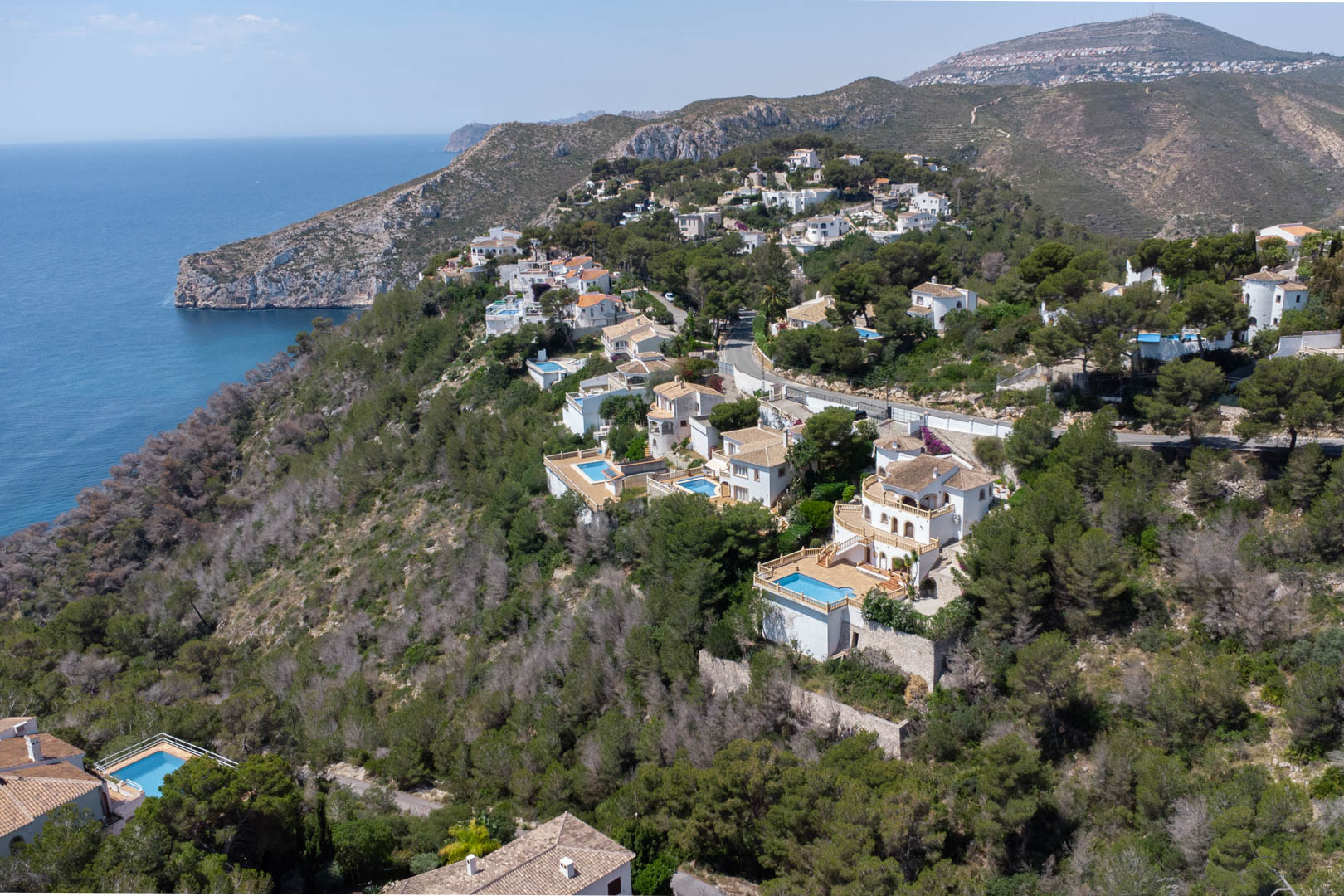 Vista aérea de la urbanización Costa Nova en Jávea, mostrando villas blancas con piscinas privadas integradas en la ladera boscosa frente al mar Mediterráneo.
