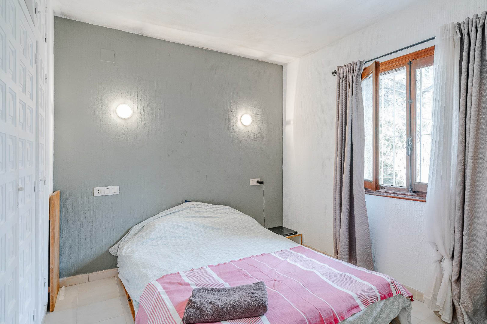 Dormitorio con pared de acento en gris, apliques de pared circulares y armario empotrado blanco. Ventana de madera con cortinas en tonos neutros y luz natural.
