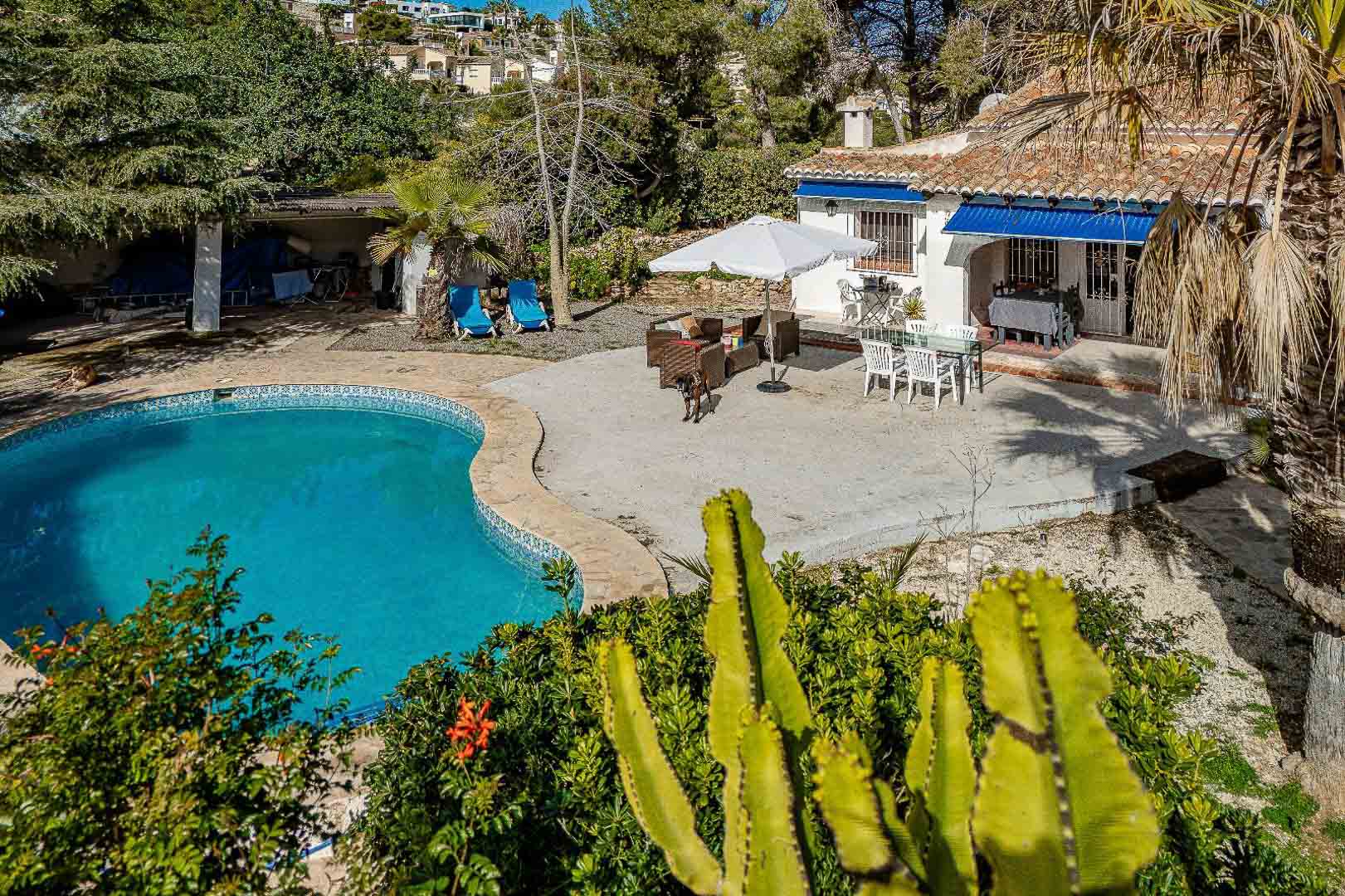 Exterior con piscina de forma irregular, amplia terraza de piedra y zona de comedor al aire libre bajo toldos azules, rodeado de vegetación mediterránea autóctona.