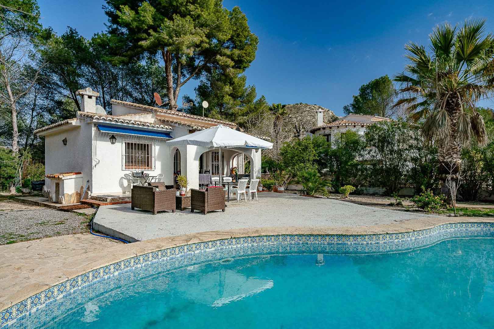 Exterior de la villa con piscina de gresite azul, amplia terraza pavimentada y porche cubierto de estilo mediterráneo rodeado de pinos y vegetación autóctona.