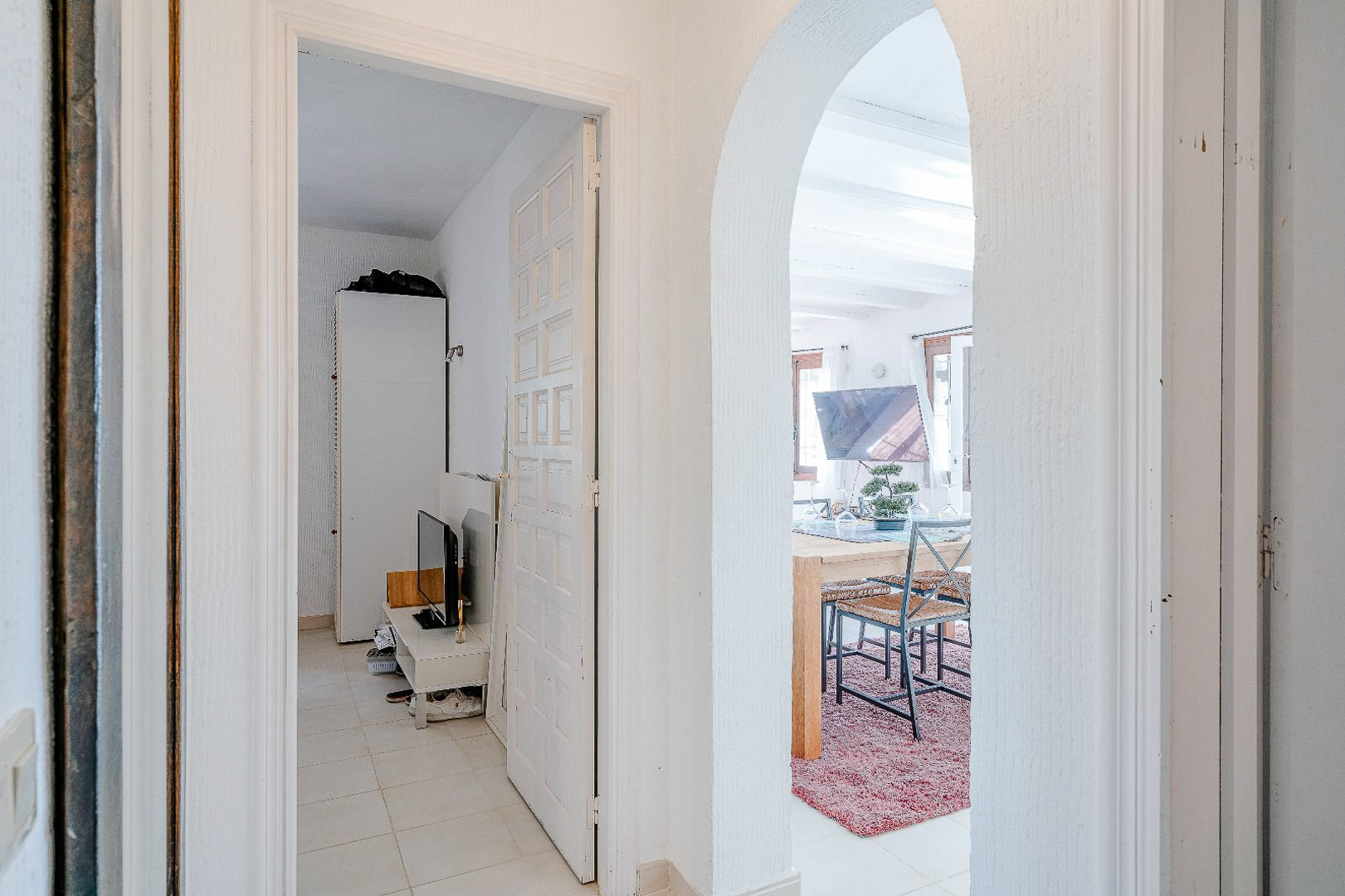 Pasillo de distribución con arco de estilo mediterráneo que conecta con el comedor y acceso a dormitorio con carpintería blanca y suelos claros.