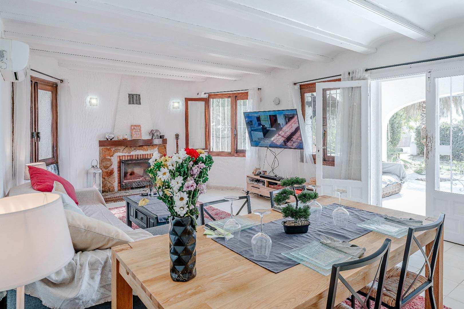 Salón-comedor con techos de vigas blancas, chimenea de piedra natural y acceso directo a la terraza exterior a través de puertas acristaladas.