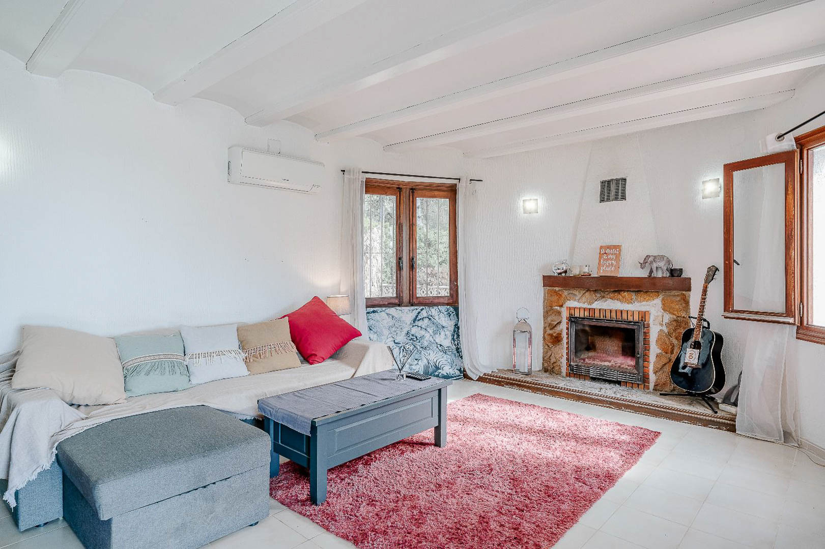 Salón luminoso con techo de vigas blancas, chimenea de piedra rústica y aire acondicionado. Los ventanales de madera aportan luz natural a este espacio de estilo mediterráneo.