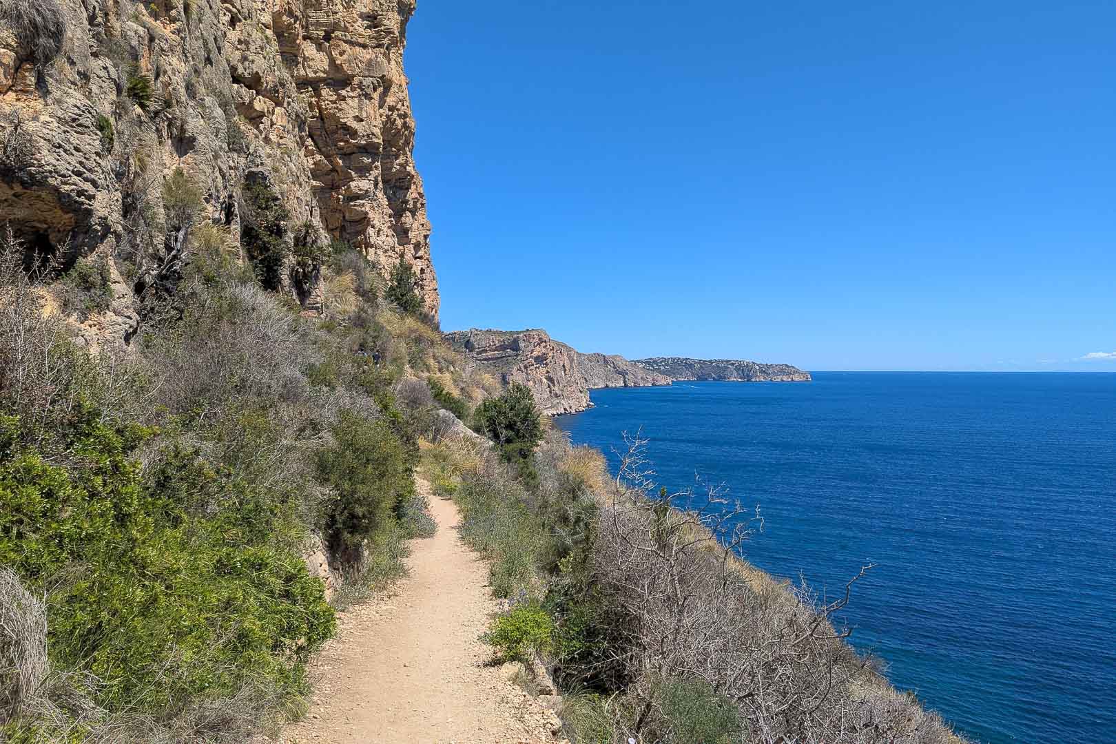 Sendero costero de tierra que bordea los acantilados de piedra caliza, ofreciendo vistas panorámicas del mar Mediterráneo bajo un cielo despejado.
