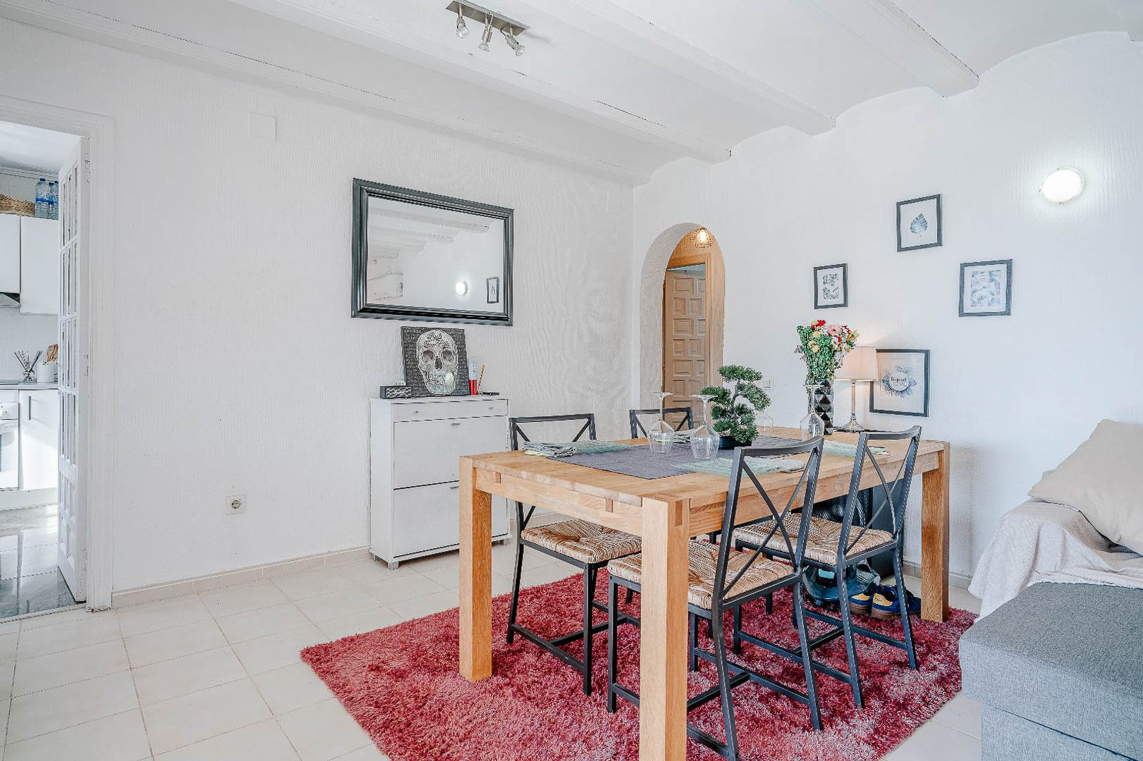 Zona de comedor con mesa de madera y sillas metálicas sobre alfombra roja, destacando techos con vigas blancas y acceso mediante arco a otras estancias.