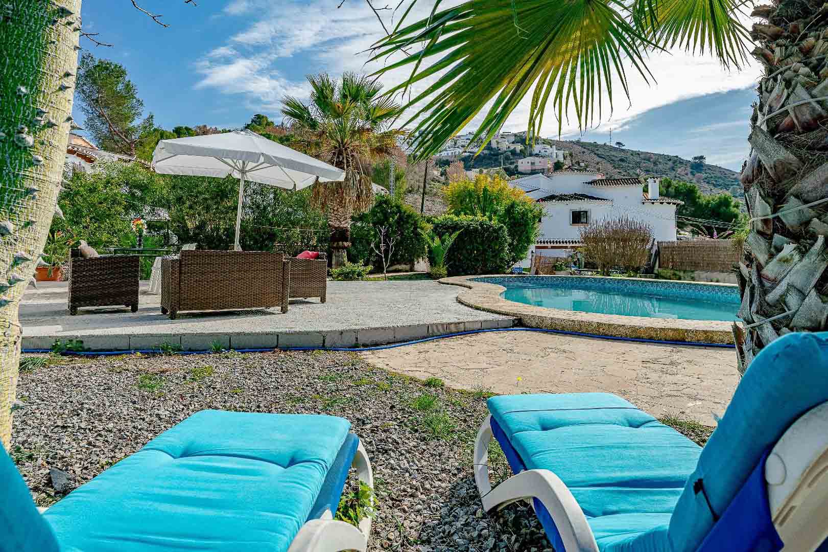 Zona de piscina con terraza de piedra y grava, equipada con tumbonas azules y mobiliario de mimbre bajo una sombrilla, rodeada de vegetación mediterránea.