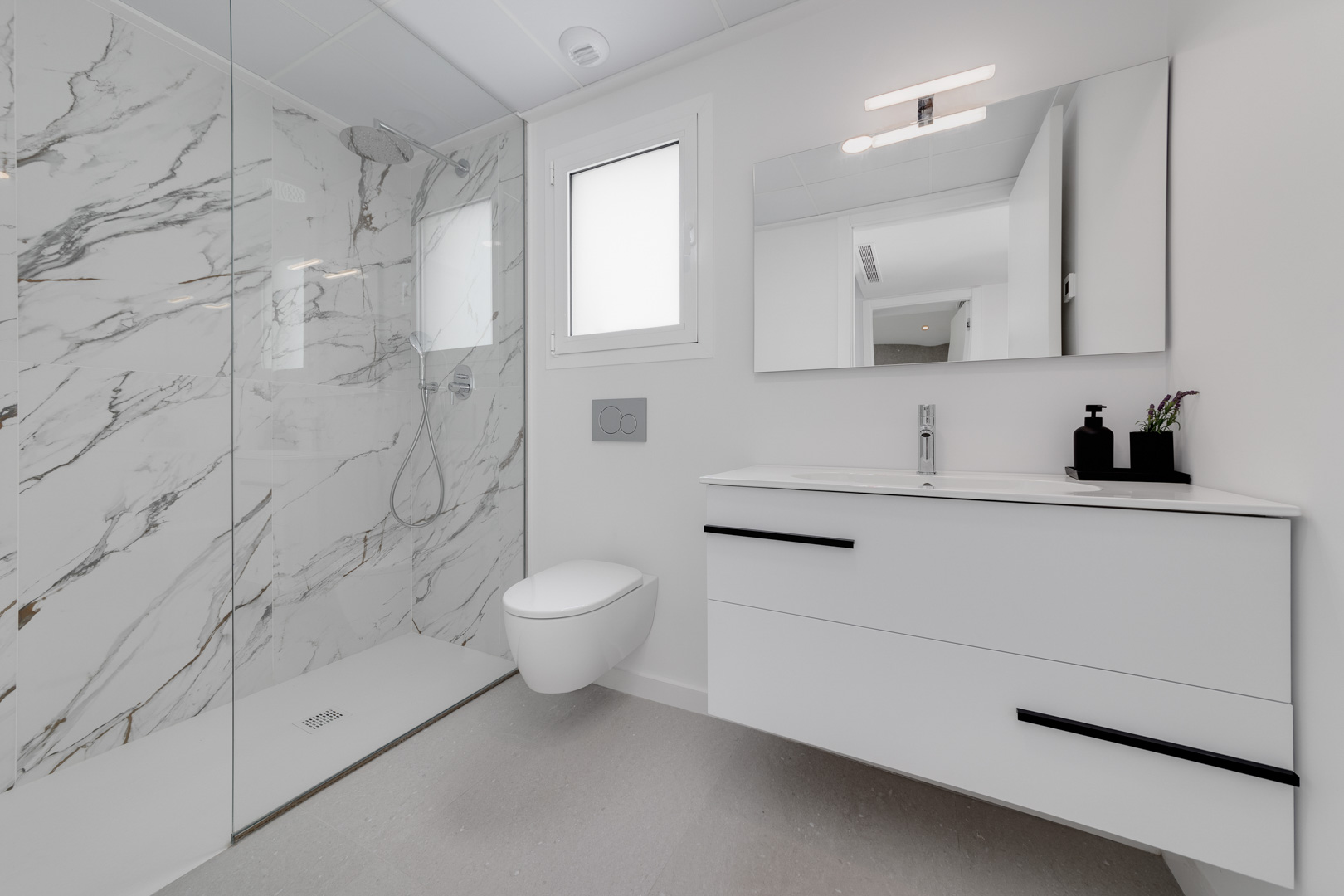 Baño contemporáneo con ducha de obra revestida en porcelánico efecto mármol, inodoro suspendido y mueble de lavabo flotante con detalles en negro.