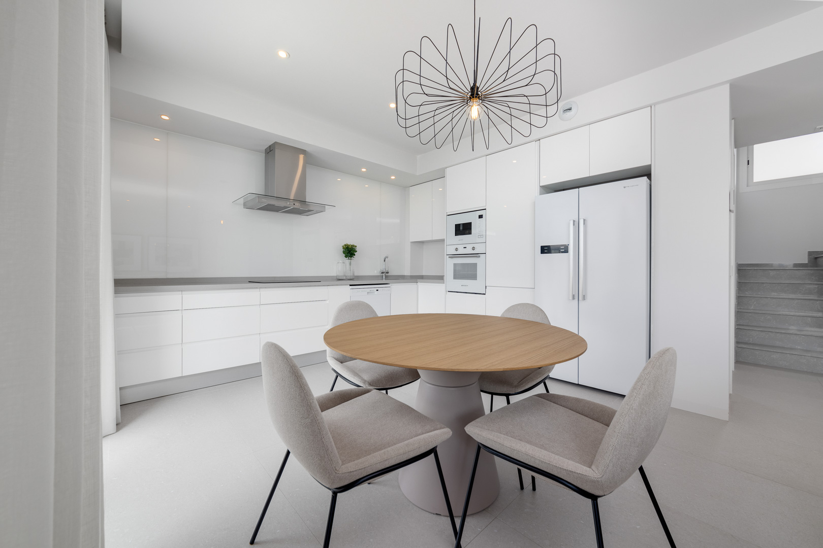 Cocina minimalista blanca con electrodomésticos integrados, encimera gris y zona de comedor con mesa circular de madera y lámpara de diseño.