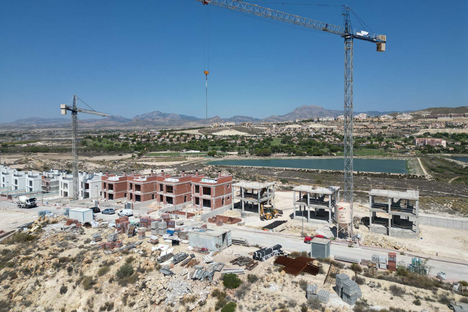Vista aérea de la fase de construcción de las villas, mostrando estructuras de ladrillo y hormigón, grúas y maquinaria con vistas al campo de golf y las montañas.