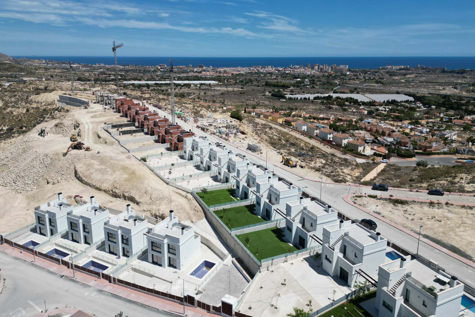 Vista aérea de villas contemporáneas de fachada blanca en una colina, con piscinas privadas y jardines orientados hacia el mar Mediterráneo y el horizonte urbano.
