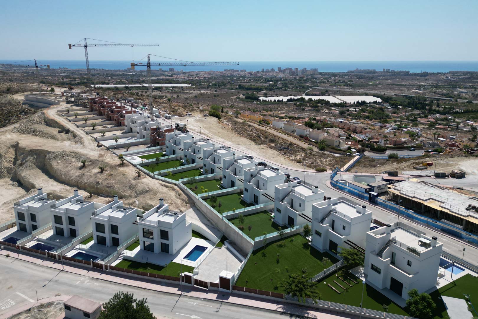 Vista aérea de villas contemporáneas en construcción sobre una colina, destacando sus fachadas blancas, jardines privados con césped y vistas despejadas al mar.