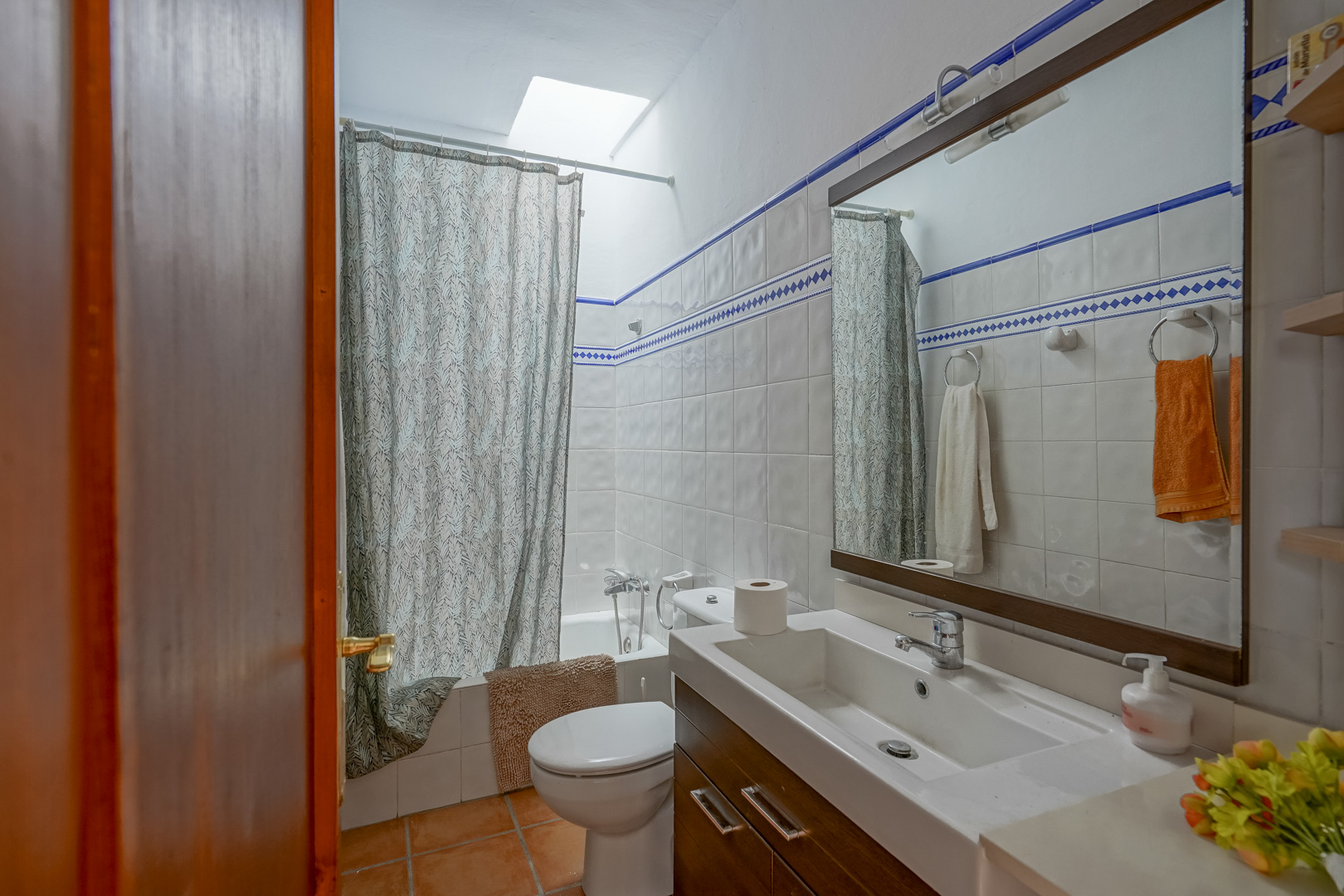 Baño funcional con azulejos blancos y cenefa azul, equipado con lavabo sobre mueble de madera, bañera con claraboya cenital y suelos de gres rústico.