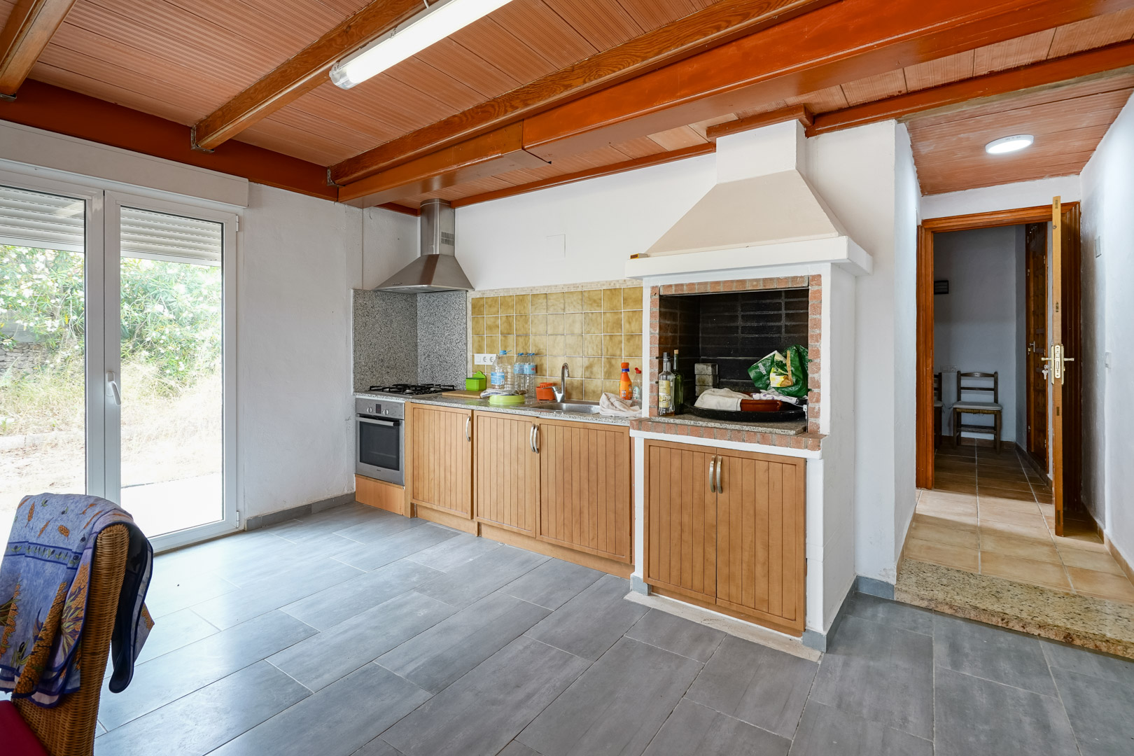 Cocina funcional con techos de madera y vigas vistas, equipada con barbacoa de obra integrada, encimeras de granito y acceso directo al exterior.