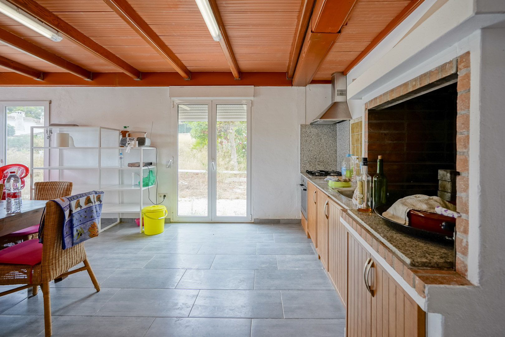Cocina rústica con techo de vigas de madera, encimera de granito y barbacoa de obra integrada, con acceso directo al exterior a través de puertas acristaladas.