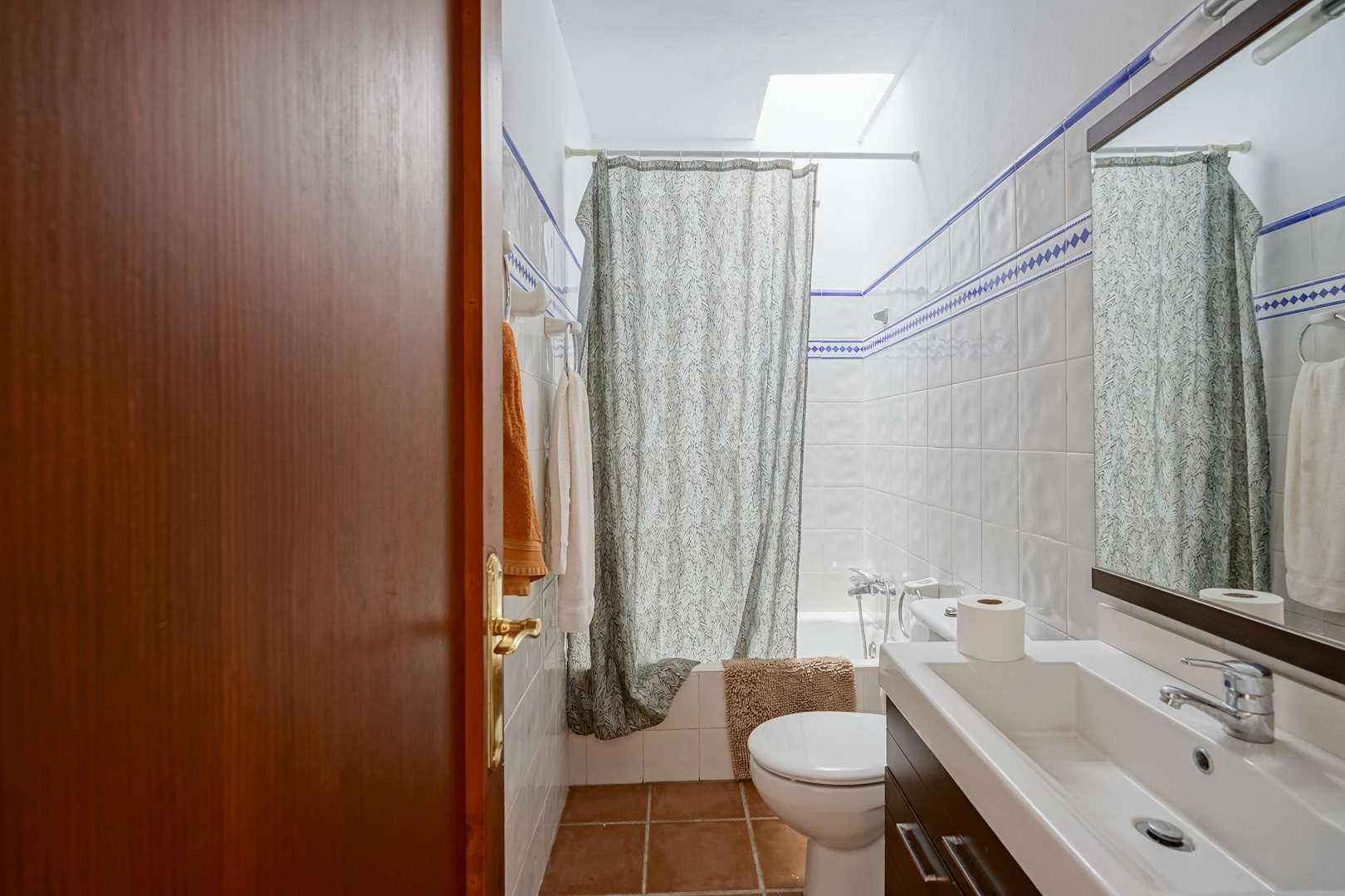 Cuarto de baño con azulejos blancos y cenefa azul, lavabo moderno sobre mueble oscuro y bañera iluminada por una claraboya cenital.