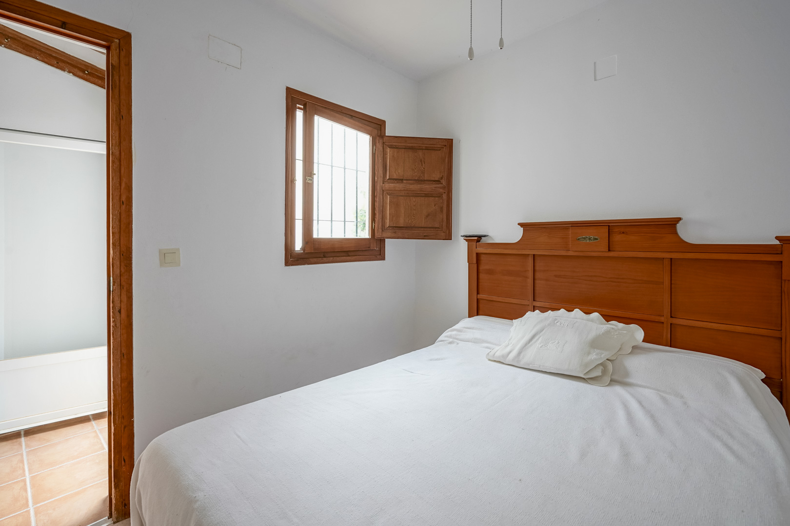 Dormitorio con cabecero de madera maciza, ventana con contraventana tradicional y acceso a estancia contigua con suelo de barro cocido.