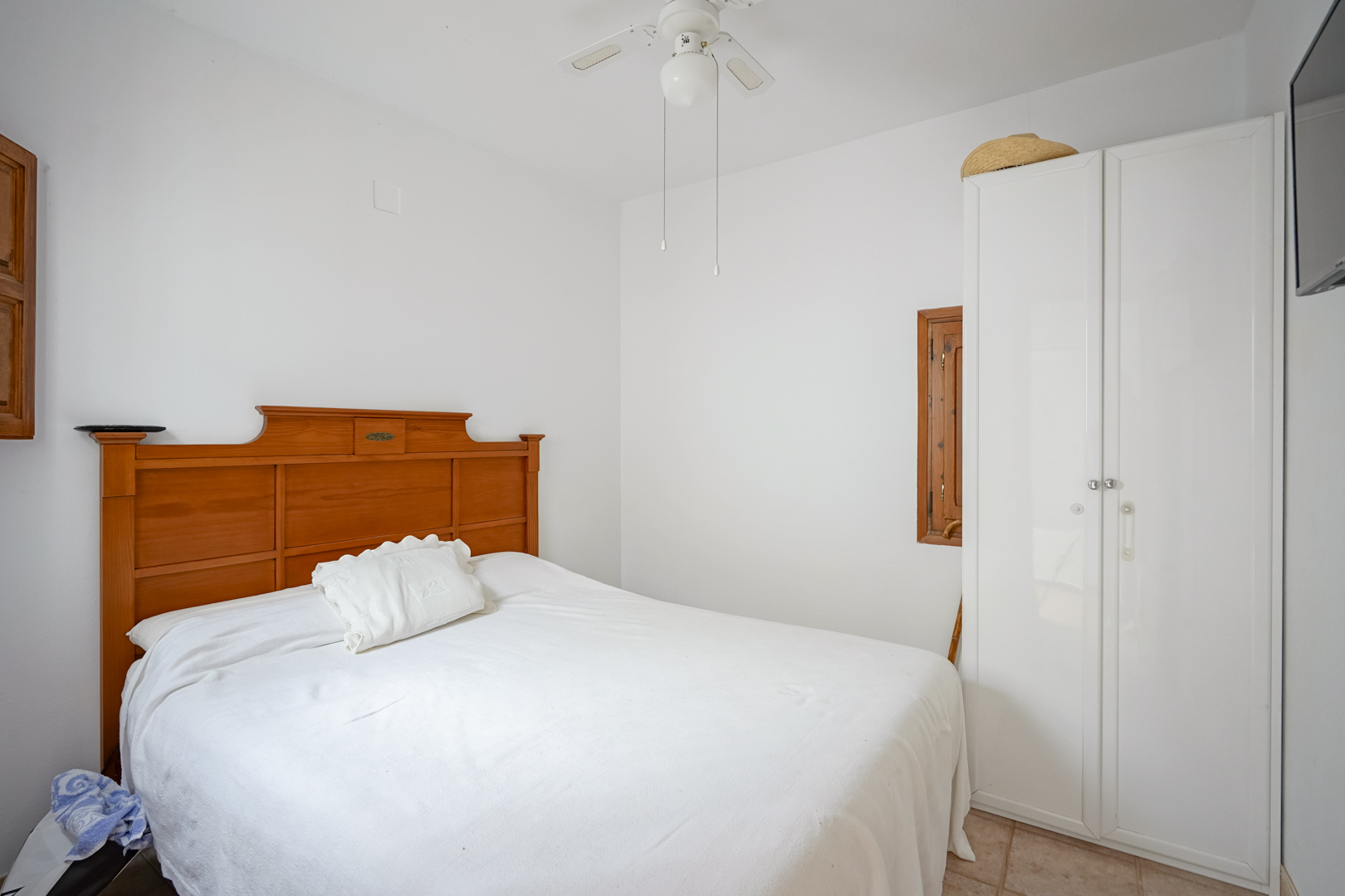 Dormitorio con cama de matrimonio, cabecero de madera, armario blanco y ventilador de techo. Paredes blancas y suelos cerámicos aportan luminosidad.