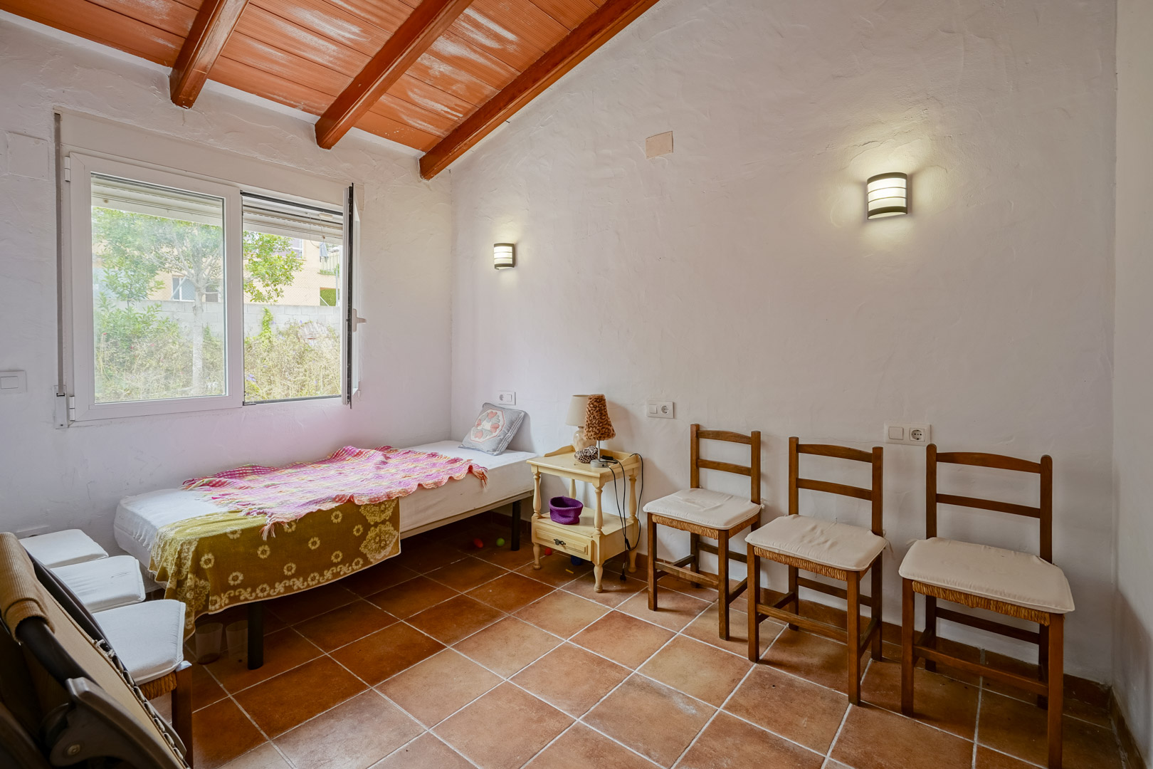 Dormitorio con techos inclinados de vigas de madera vistas, suelos de terracota y paredes blancas. Dispone de iluminación mediante apliques de pared y ventana al exterior.